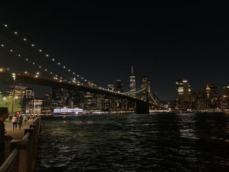 people are standing on the edge of a pier looking at the city lights and bridge