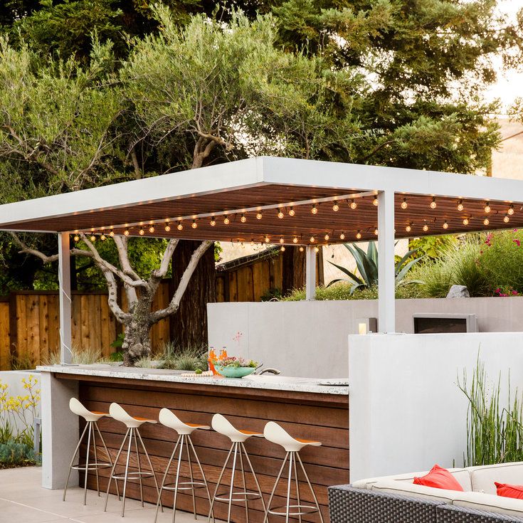 an outdoor kitchen with bar stools and lights on the outside wall, next to trees