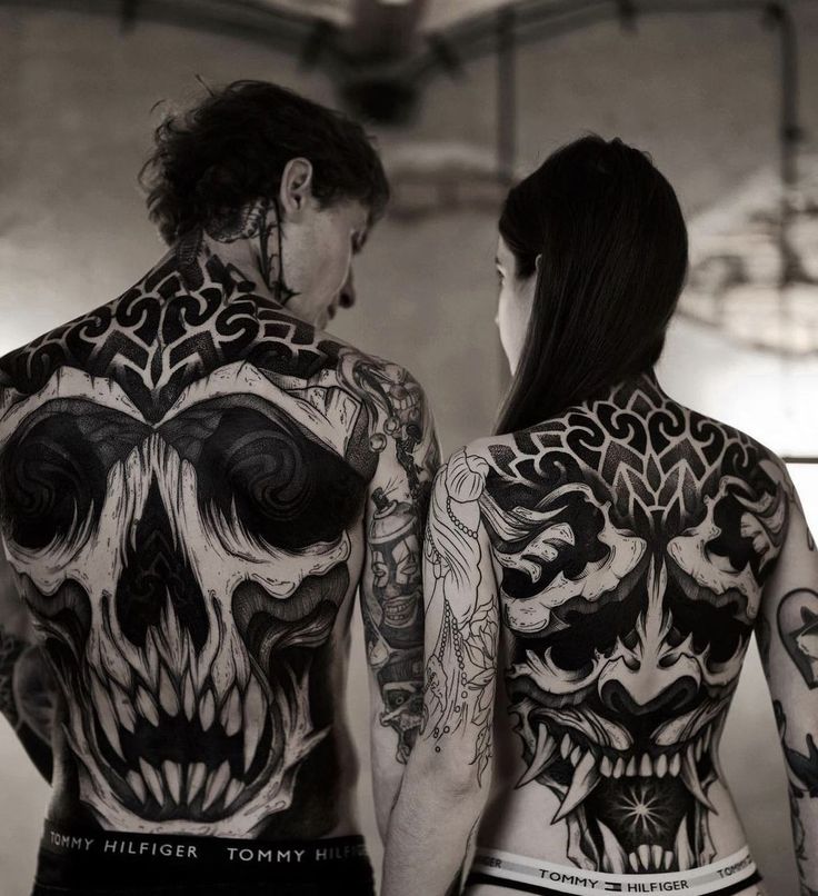 two people with tattoos on their back standing next to each other and looking at each other