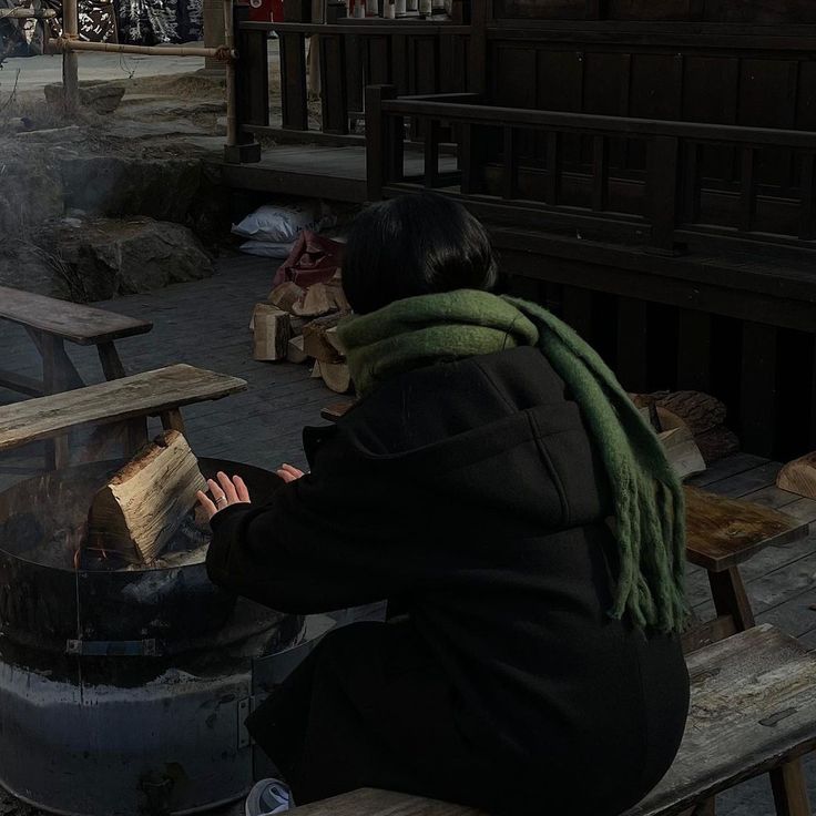 a woman sitting on a bench next to an open fire pit with logs in it