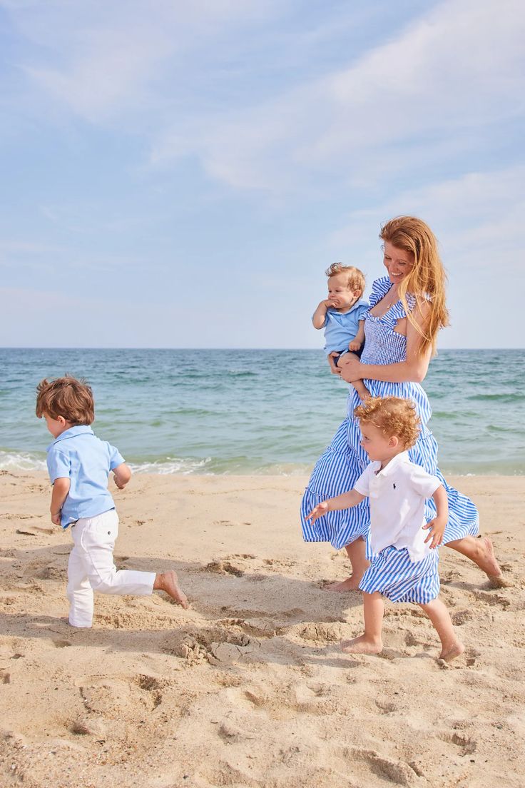 a woman in a blue dress holding a baby on the beach with three other children