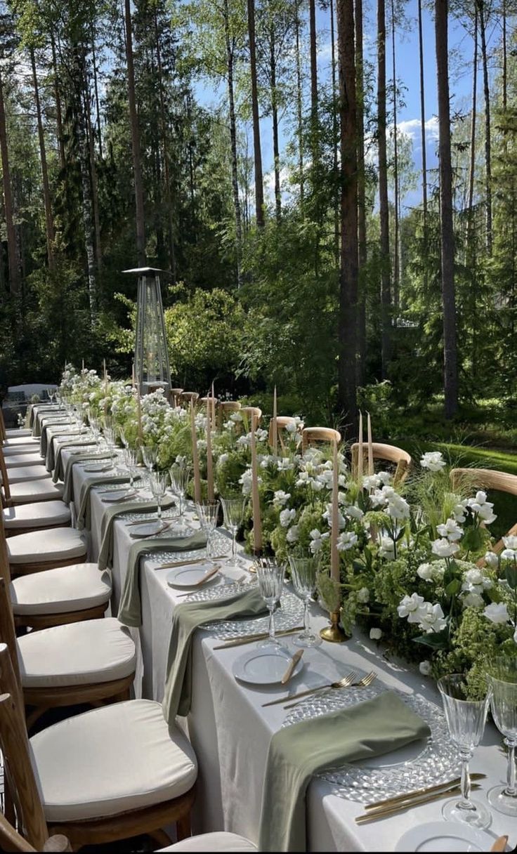 a long table is set with white flowers and place settings for an outdoor dinner in the woods