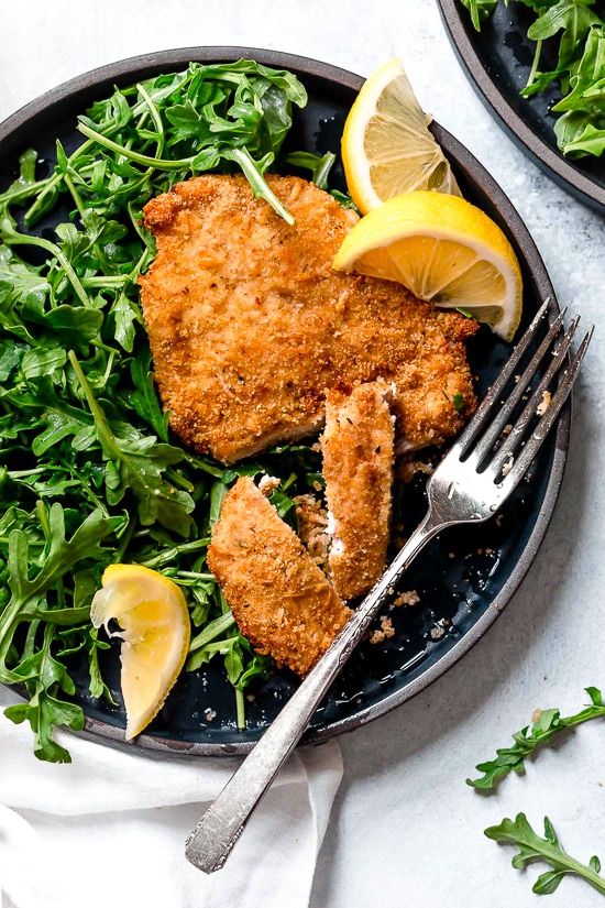 two pieces of chicken on a plate with greens and lemon wedges