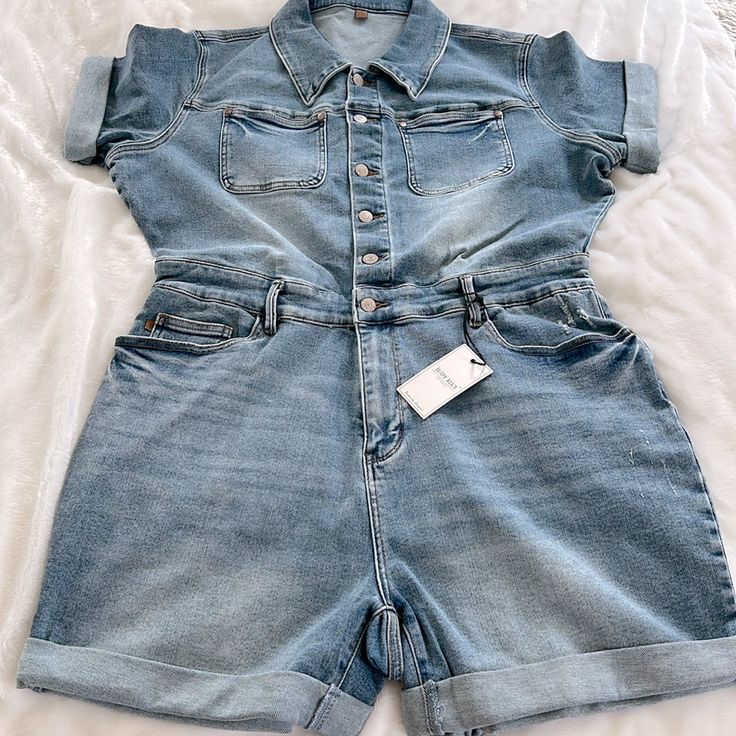 This Judy Blue Jumper Is A Light-Medium Wash Denim With Buttons Down The Top, A Stretchy Band Across The Waist And A Zipper Fly. The Arm And Short Hems Are Cuffed, And There Are Pockets On The Chest And Front And Back Of The Shorts. The 2x Measures About 24” From Underarm To Underarm, About 19” Across The Waist, And Has An Inseam Of About 5”. The 3x Measures About 25” From Underarm To Underarm, About 20” Across The Waistband, And Has An Inseam Of About 5.5”. B2 Medium Wash Denim Shortalls With Relaxed Fit, Relaxed Fit Denim Shortalls In Medium Wash, Medium Wash Denim Shortalls In Relaxed Fit, High Rise Denim Shortalls For Spring, Blue Relaxed Fit Denim Shortalls, Denim Shortalls For Spring, Dark Wash High Rise Denim Shortalls, High Rise Dark Wash Denim Shortalls, Fitted Medium Wash Shortalls For Spring