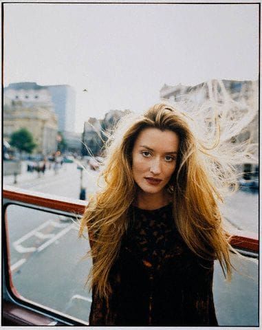 a woman with long blonde hair standing in front of a bus on the street and looking at the camera