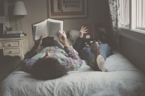 two people are laying on a bed and reading books in front of a window with curtains