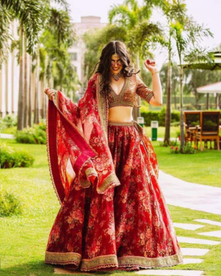 a woman in a red and gold lehenga dancing on the grass with palm trees behind her