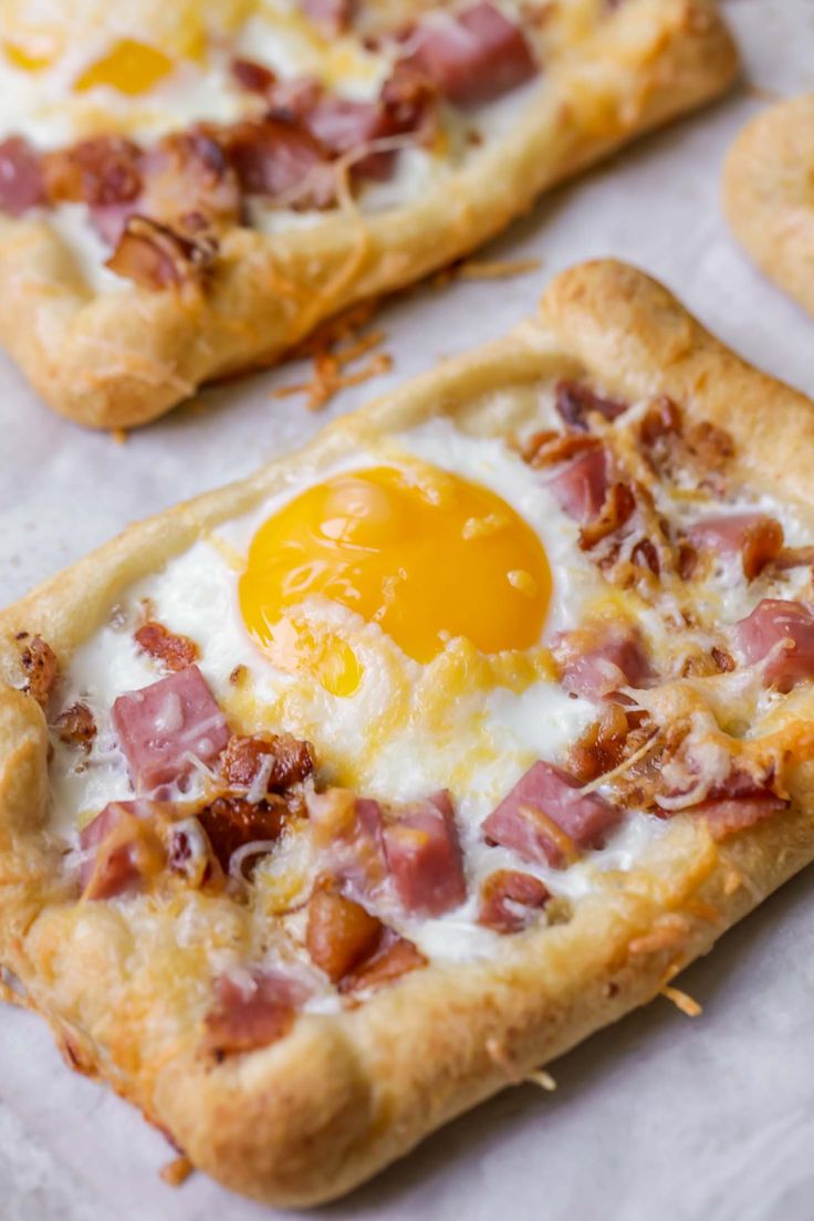 two square pizzas with bacon and an egg on top are sitting on wax paper