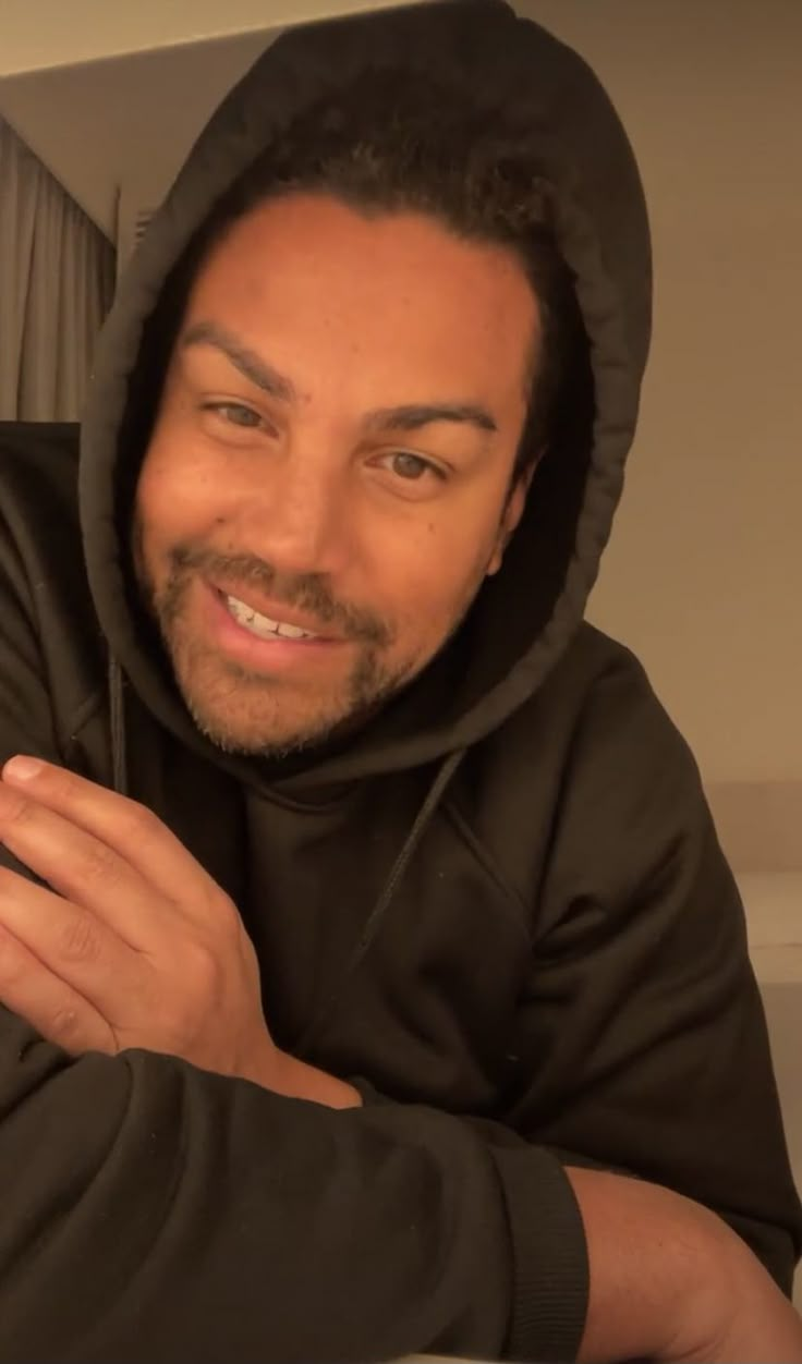 a man in a black hoodie is smiling and holding his hands on his chest