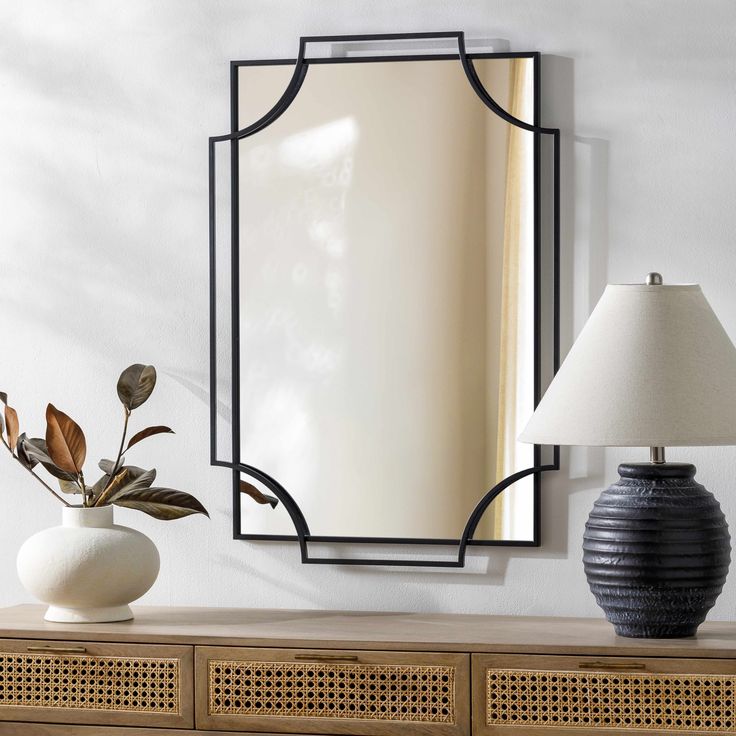 a mirror on the wall above a dresser with a vase and lamp next to it