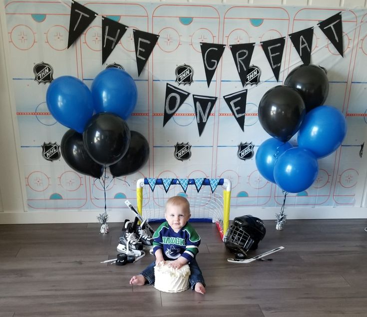 a baby boy sitting on the floor in front of balloons and hockey gear, with some black and blue streamers