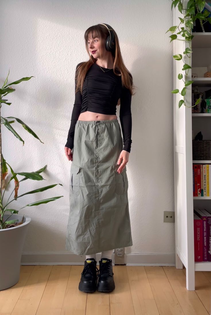 an outfit with a cargo skirt, chunky black boots, crop top & headphones How To Style Cargo Skirt Women, Cargo Skirt Maxi Outfit, Cargos Skirts Outfits, Cargo Skirt Inspo Outfit, Cargo Long Skirts, Cargo Skirt Green Outfit, Green Cargo Maxi Skirt Outfit, Aesthetic Cargo Skirt Outfit, Green Cargo Maxi Skirt