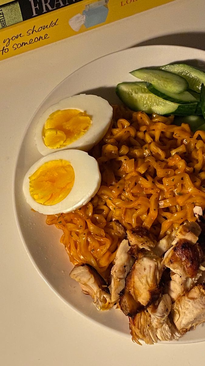 a white plate topped with noodles, chicken and hard boiled eggs next to cucumbers