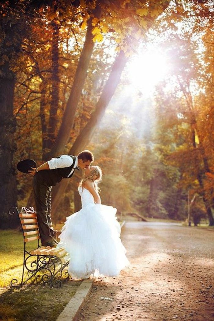 a bride and groom kissing on a bench in the middle of an autumn park setting