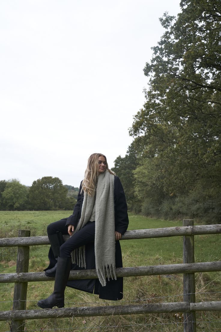 a woman sitting on top of a wooden fence next to a lush green field with trees