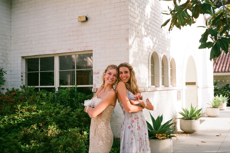 two women standing next to each other in front of a white brick building with plants