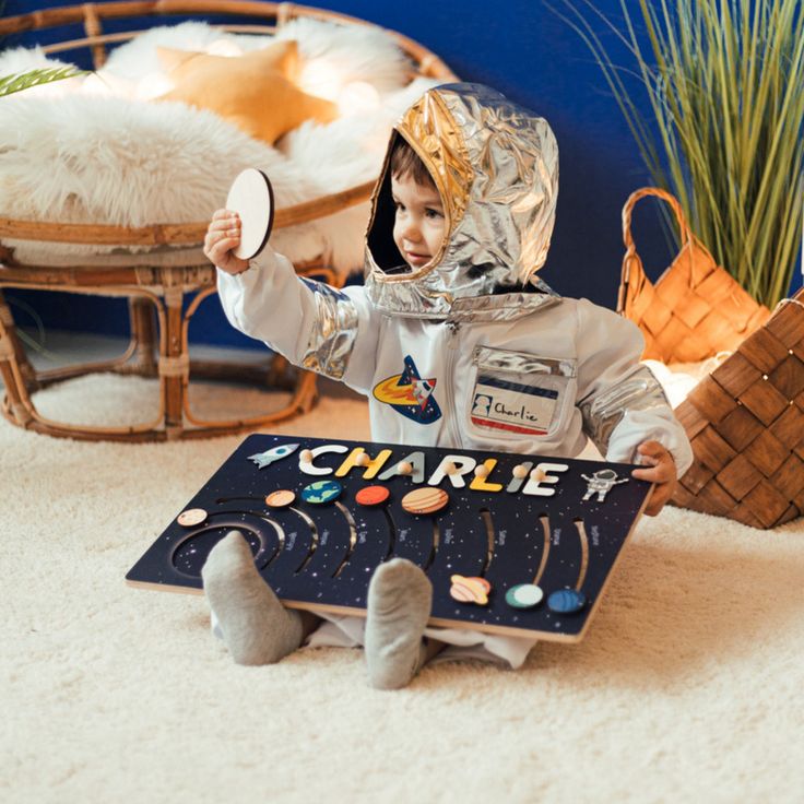 a small child in an astronaut suit holding a sign that says charlie on it and sitting on the floor