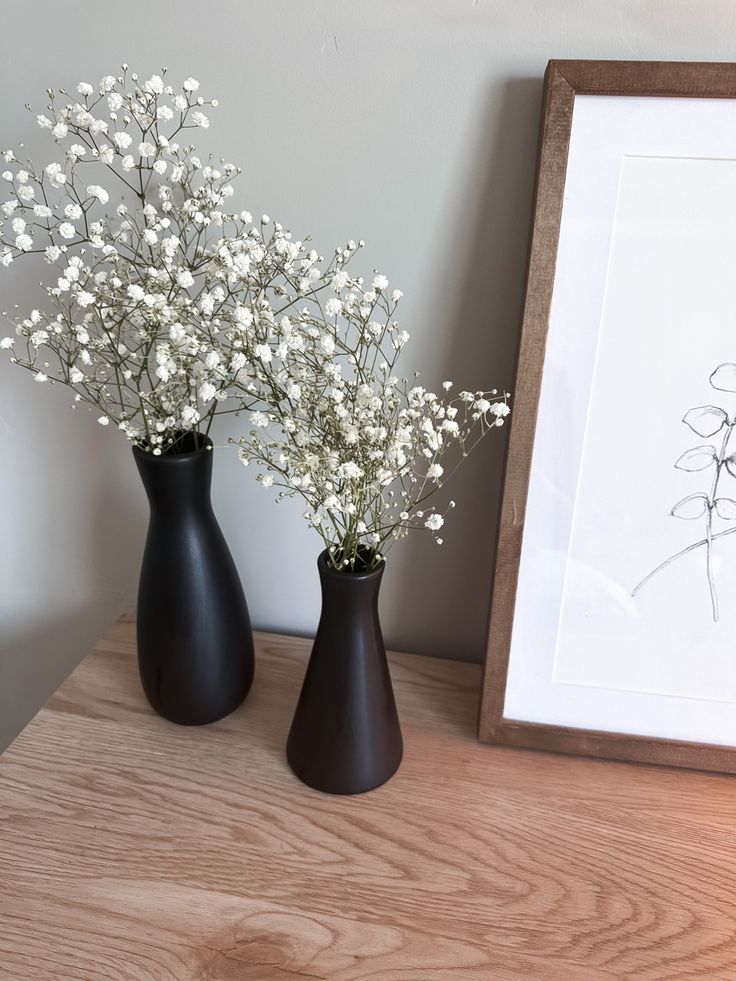 two black vases with white flowers in them on a table next to a framed drawing