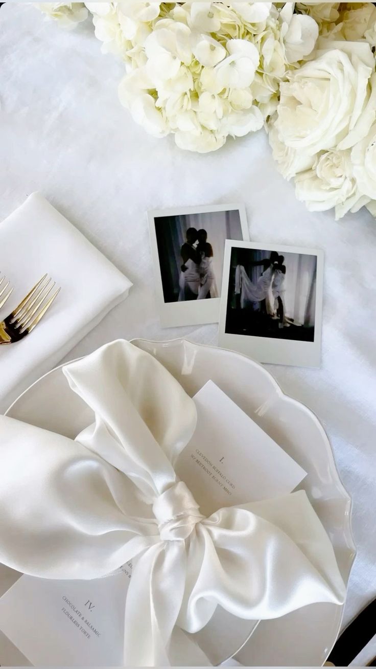 the table is set with white flowers and photos on it, along with two gold - plated utensils