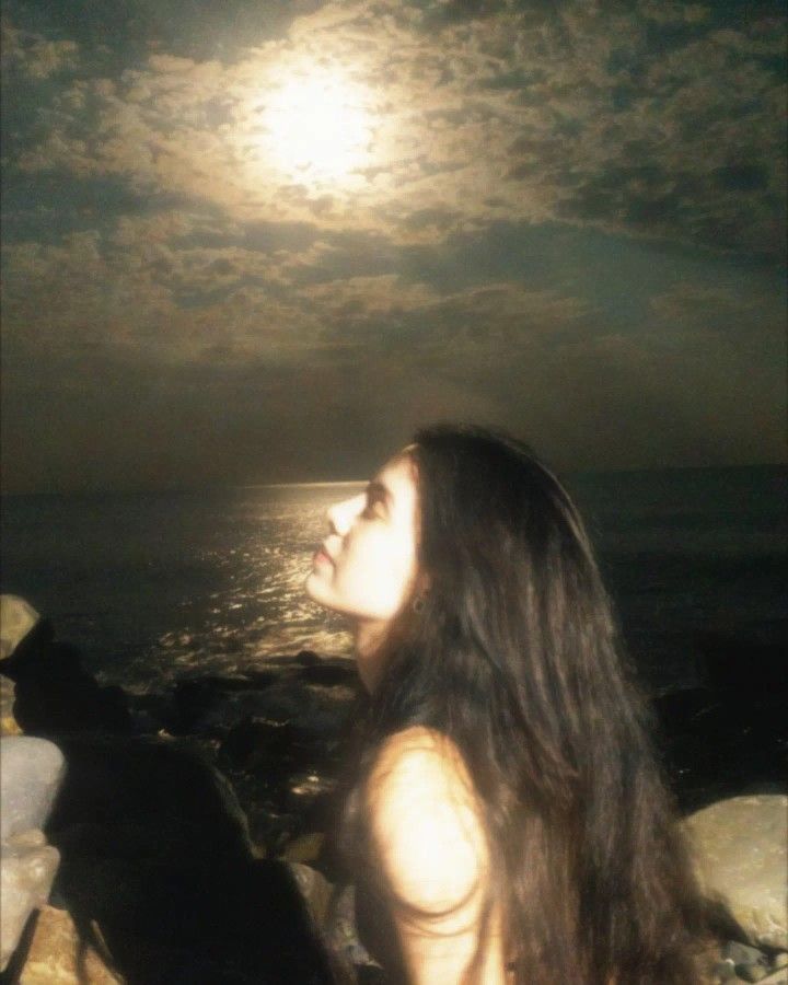 a woman sitting on top of a rock next to the ocean under a full moon