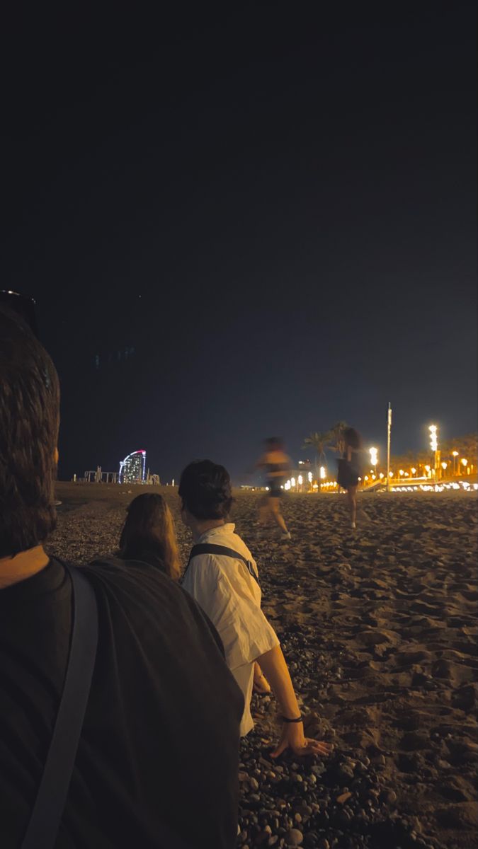 two people sitting on the beach at night looking out over the water and lights in the distance