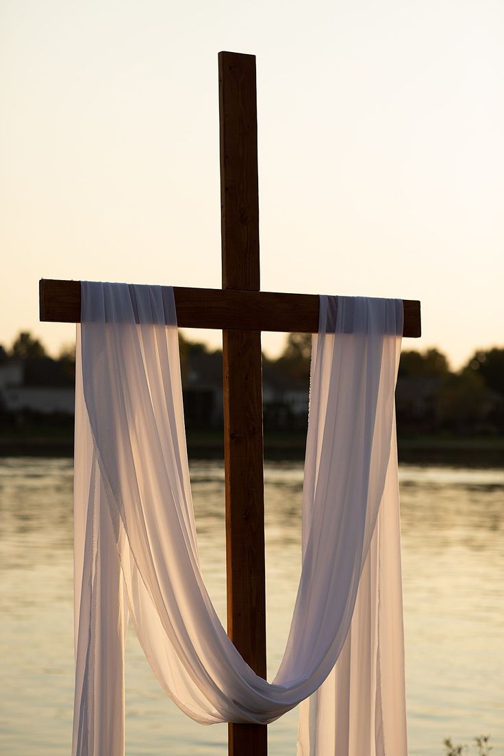 a cross with sheer curtains hanging from it's sides near the water at sunset