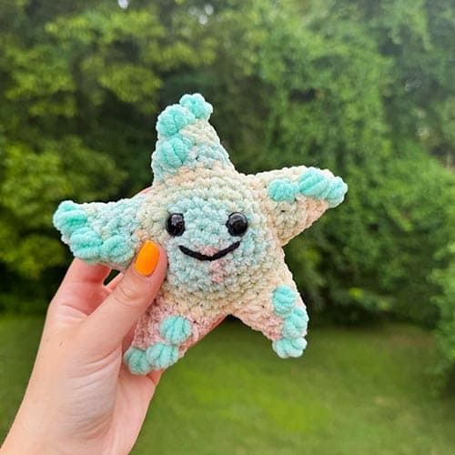 a hand holding a small crocheted starfish toy in front of some trees