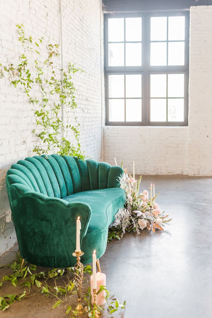 a green velvet couch sitting in front of a window next to candles and greenery