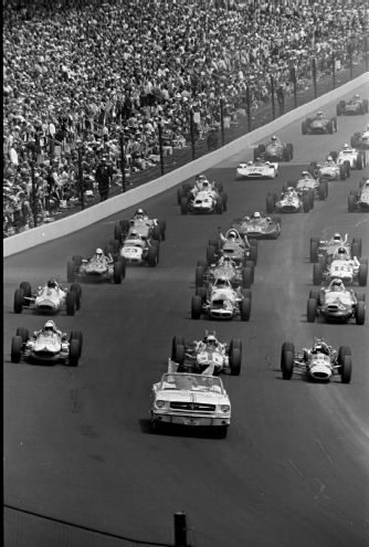 a group of cars driving down a race track with people watching from the sidelines