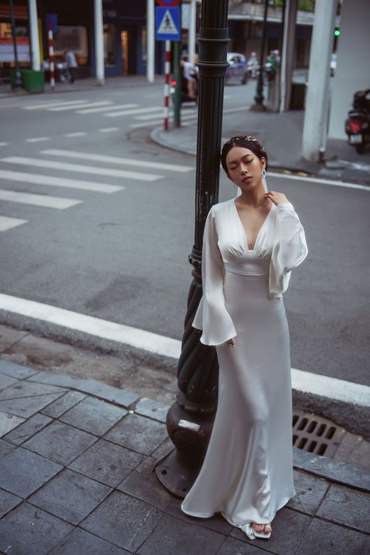 a woman standing next to a lamp post on the side of a road wearing a white dress