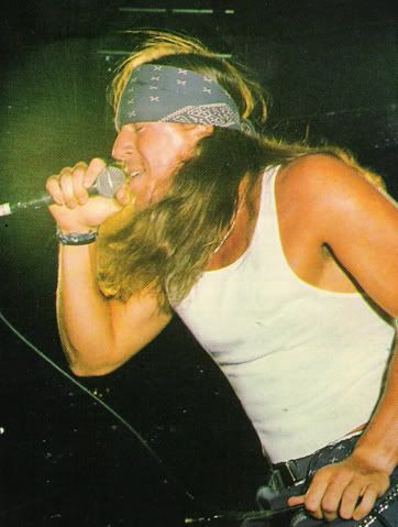 a man with long hair and bandana on holding a microphone to his mouth while standing in front of a stage