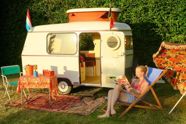 a woman sitting in a lawn chair next to an old camper with the door open