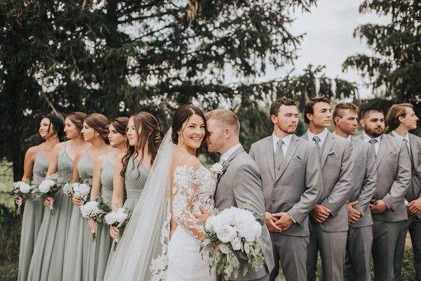 a group of people standing next to each other in front of trees and grass with one woman wearing a wedding dress