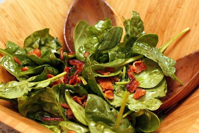 a wooden bowl filled with spinach and bacon on top of a table next to a spoon