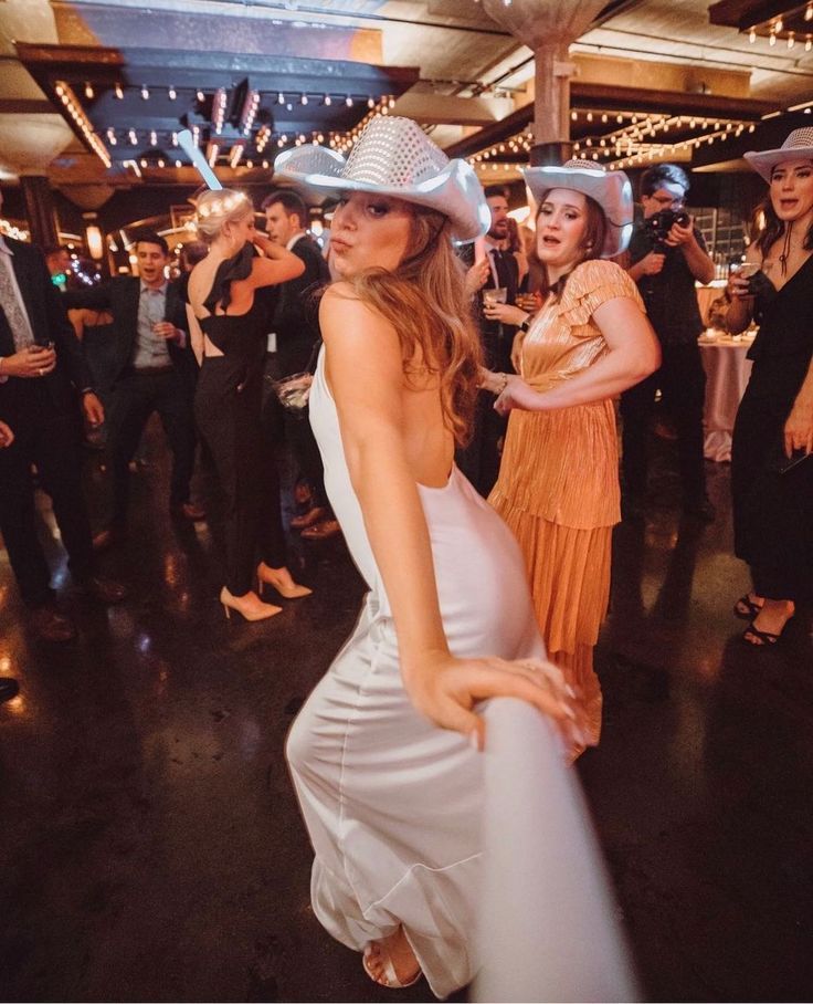 two women in white dresses and cowboy hats dancing at a party with people standing around