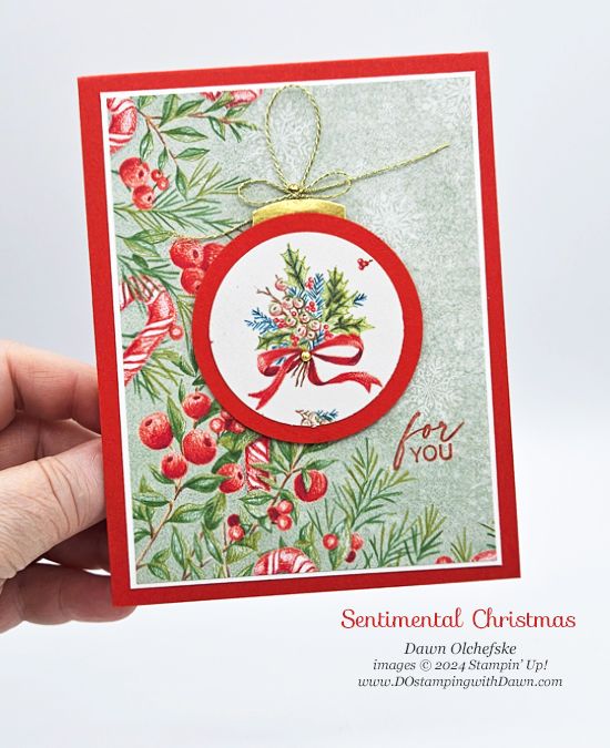 a hand holding up a christmas card with a red ribbon and holly decoration on it