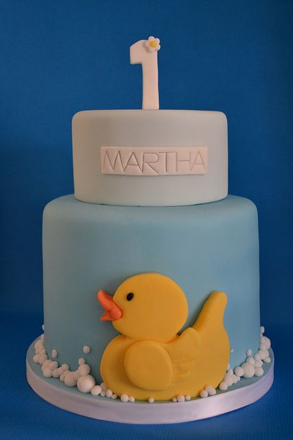 a blue and white cake with a rubber ducky on the front, first birthday candle in the background