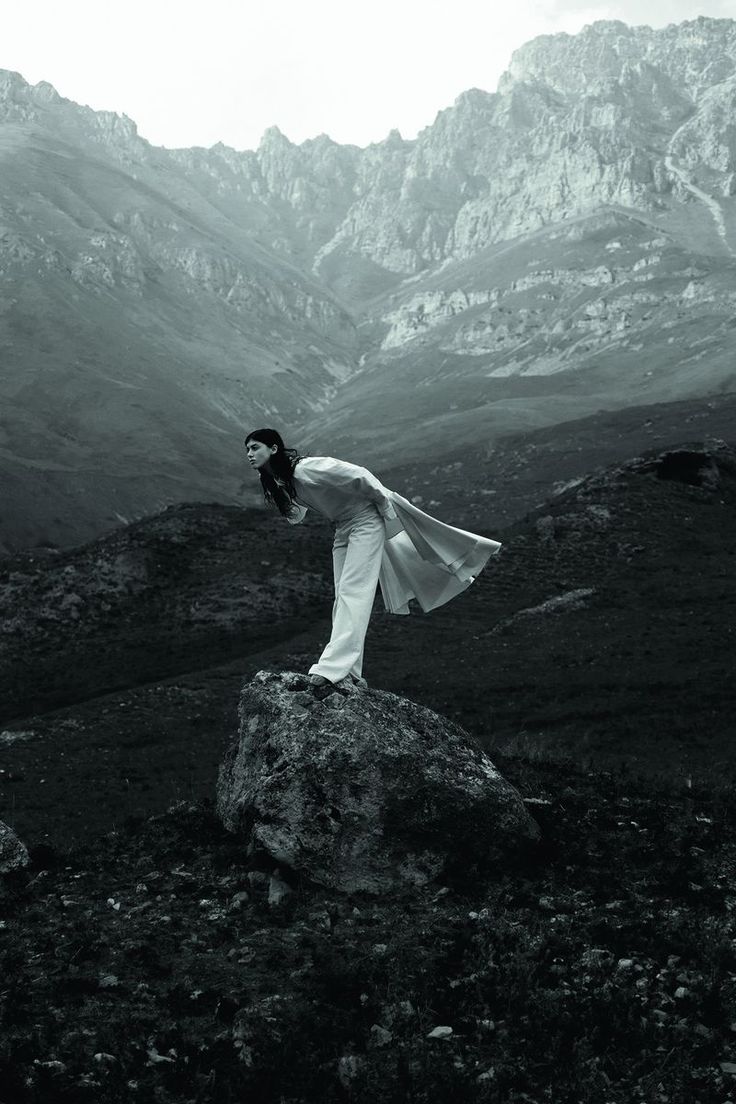 a woman standing on top of a large rock in the middle of a mountain range