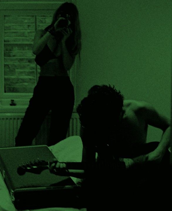 two people in a dark room, one playing guitar and the other on a laptop