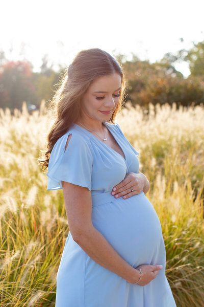 a pregnant woman in a blue dress standing in tall grass with her hands on her belly