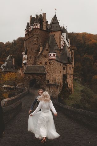 a bride and groom walking on a bridge in front of an old castle with fall foliage