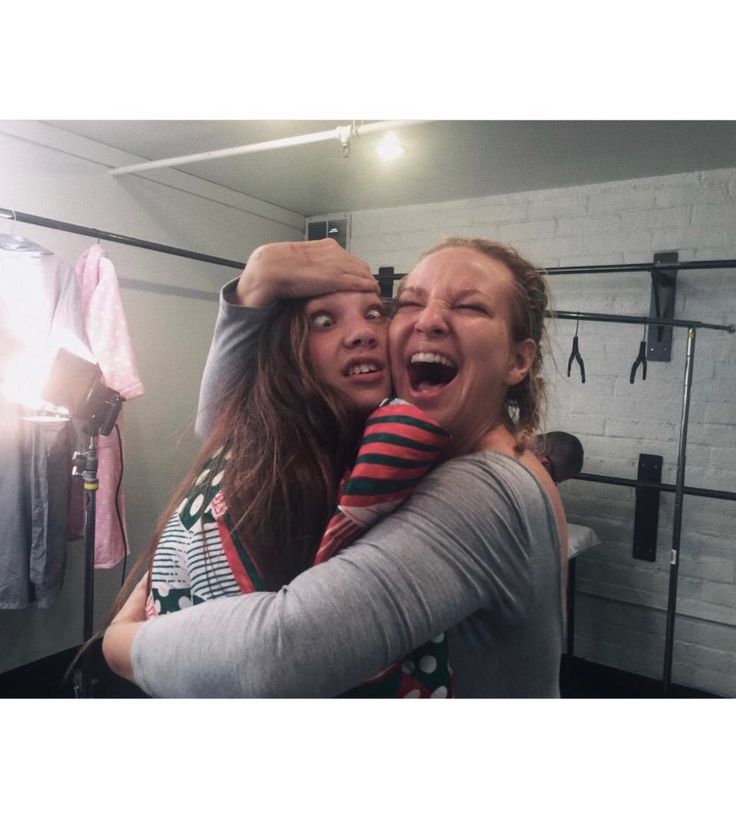 two women hugging each other in front of a mirror with clothes hanging on the wall behind them