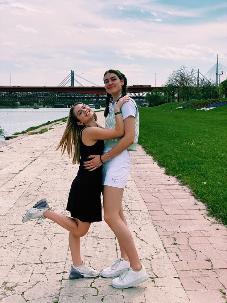 two young women standing next to each other in front of a body of water with a bridge in the background