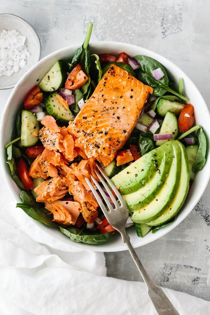 a salad with salmon, avocado and tomatoes in it on a white table