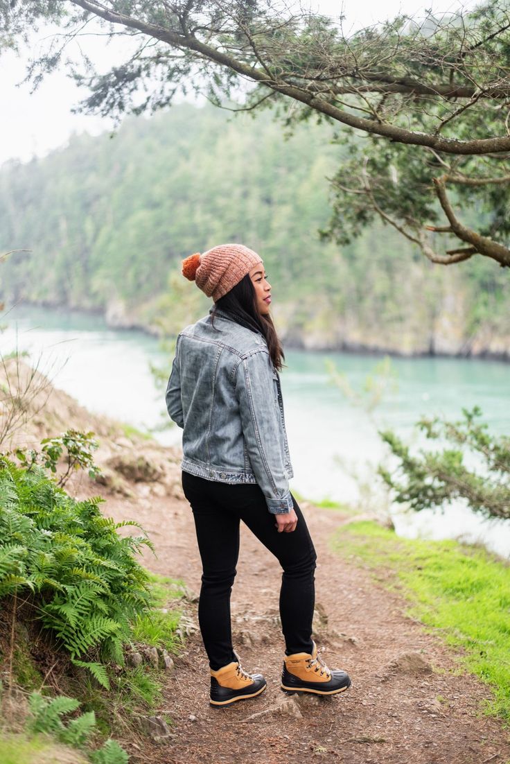 a woman standing on a dirt path next to a river wearing a denim jacket and beanie