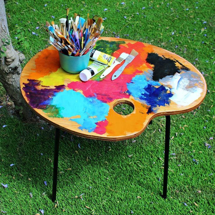 a colorful table with paint and brushes on it in the grass next to a tree