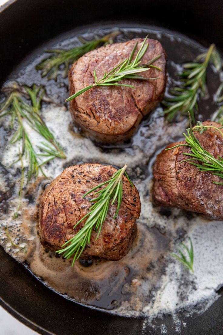 three steaks cooking in a skillet with some rosemary sprigs on top