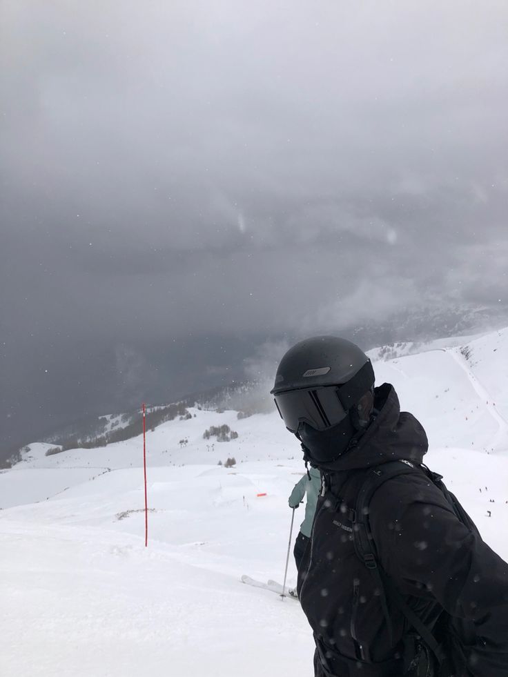 a man in black jacket and helmet standing on snow covered slope with skis under cloudy sky