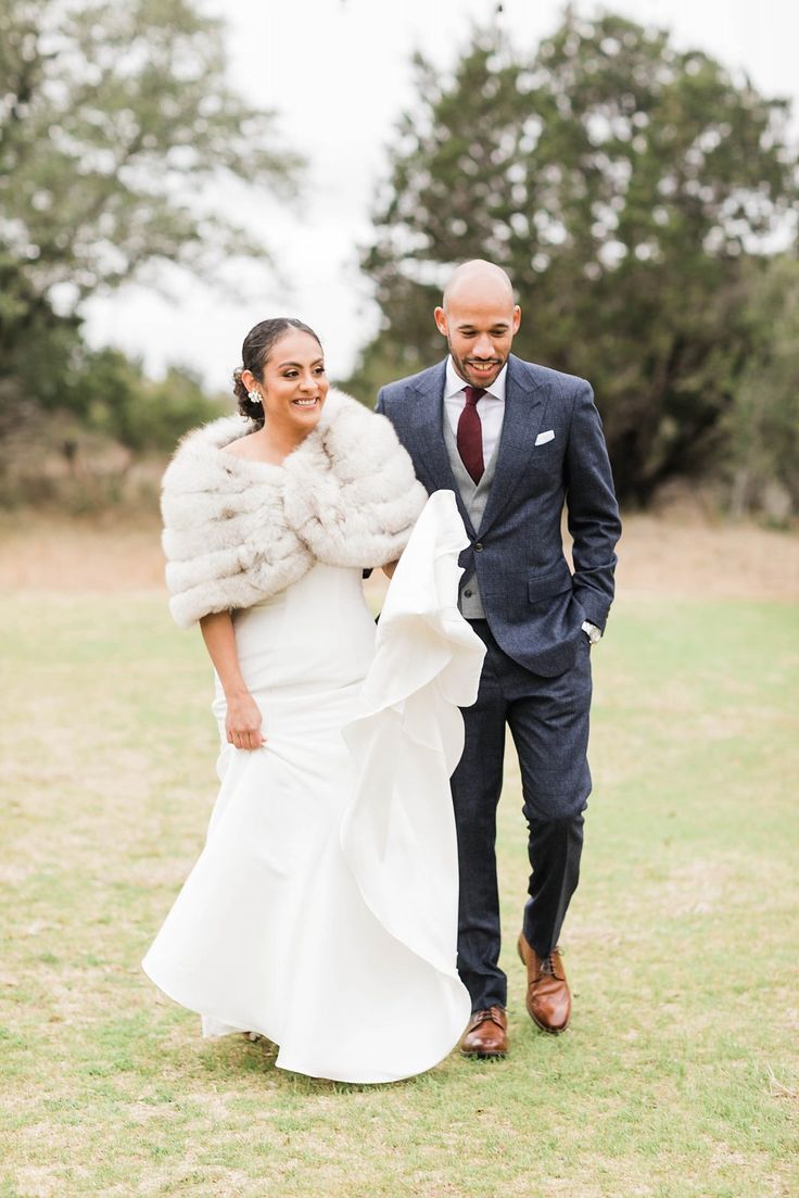 a bride and groom walking through the grass in their wedding attire with fur stolers