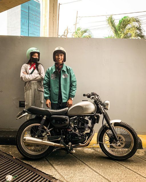 two people standing next to a motorcycle parked on the side of a road in front of a wall